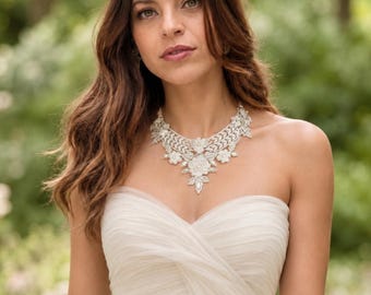 May include: A statement necklace featuring intricate silver-toned metalwork, clear crystals, and white floral-shaped beads. The design includes layered leaf patterns and a central cluster of crystals, creating a wide, ornate collar shape that rests against the collarbone.