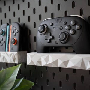 May include: A black Nintendo Switch Pro controller and a pair of Joy-Con controllers resting on white, geometric, faceted wall-mounted shelves. A small white holder contains several game cartridges, all mounted on a black pegboard wall panel.