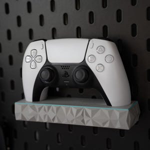 May include: A white and black wireless game controller resting on a light gray rectangular shelf with a geometric faceted front surface. The controller features a central touchpad, two analog sticks, and standard directional and action buttons.
