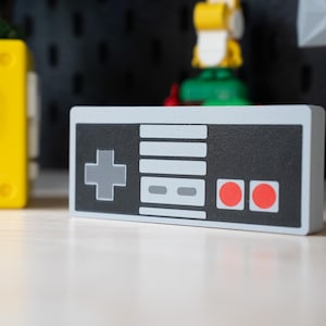 May include: A rectangular wooden block shaped like a retro video game controller. The front features a black panel with a gray directional pad, a central gray button area, and two red circular buttons. The edges of the block are light gray.