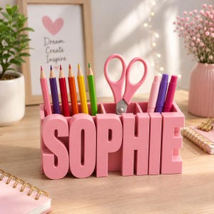 Puede incluir: Un organizador de escritorio rosa con el nombre SOPHIE en letras grandes y en relieve en la parte frontal. El organizador tiene varios compartimentos que contienen lápices de colores, unas tijeras rosas y dos rotuladores rosa y morado.