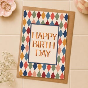 May include: A rectangular greeting card featuring a colourful harlequin diamond pattern in shades of blue, red, yellow, and green. The centre contains a white rectangular panel with the text HAPPY BIRTH DAY in bold, orange, block letters.