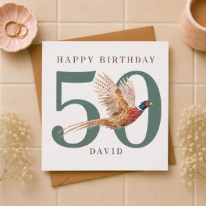 Peut inclure: Une carte de vœux d'anniversaire carrée avec le texte HAPPY BIRTHDAY en haut et DAVID en bas en police marron foncé. Un grand nombre 50 vert est imprimé au centre avec une illustration à l'aquarelle d'un faisan volant à travers celui-ci.