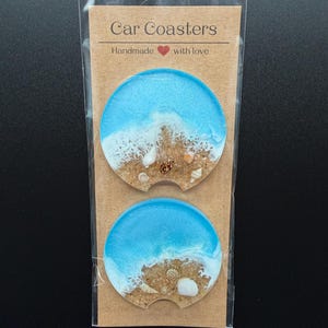 May include: Two circular resin car coasters featuring a beach scene with blue water, white foam, and tan sand embedded with small seashells. The coasters are packaged in a clear plastic sleeve on a brown card labeled Car Coasters Handmade with love.
