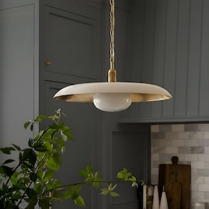 May include: A pendant light fitting featuring a wide, shallow, off-white saucer-shaped shade with a metallic gold interior finish. A central, spherical white glass globe is suspended beneath the shade, connected by a brass-toned metal chain.