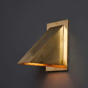 May include: A wall-mounted light fixture featuring a rectangular backplate and a triangular, wedge-shaped brass shade. The metal surface has a brushed, metallic finish, and the open bottom edge emits a warm, downward-facing glow of light.