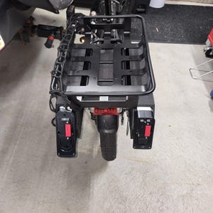 May include: A black cargo carrier with a rectangular basket and a rear light assembly. The carrier is mounted on a vehicle and includes side lights with red accents. The design is functional and robust, suitable for transporting goods.