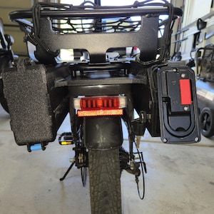 May include: Rear view of a black electric bicycle with a cargo rack and panniers. The bike has a red taillight and a black mudguard. The tires are black with a textured tread. A black storage case is attached to the side.