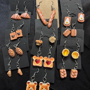 May include: A collection of miniature food-themed dangle earrings displayed on black and brown cardboard cards. The earrings feature various baked goods and breakfast items including croissants, baguettes, toast with hearts, fried eggs, and bear-shaped bread.