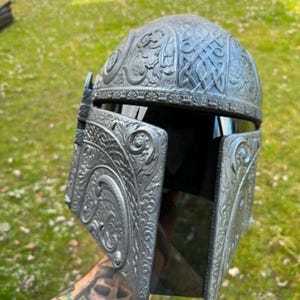 May include: A silver-grey helmet with detailed Celtic knot and floral patterns. The helmet has a dark visor. The design suggests a fantasy or sci-fi theme, possibly for cosplay or display. The helmet's design is reminiscent of historical or fictional armor.