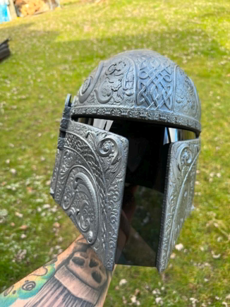 May include: A silver-grey helmet with detailed Celtic knot and floral patterns. The helmet has a dark visor. The design suggests a fantasy or sci-fi theme, possibly for cosplay or display. The helmet's design is reminiscent of historical or fictional armor.