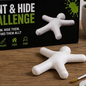 May include: A Paint & Hide Challenge box with white text and green accents, featuring two blank white humanoid figures. The box displays the text: PAINT & HIDE CHALLENGE. PAINT THEM. HIDE THEM. CAN YOU FIND THEM ALL? A paintbrush and paint palette are visible.