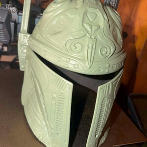 May include: A light green Mandalorian helmet with detailed features, including a T-shaped visor and antenna. The helmet is made of a robust material and has a complex design with various shapes and patterns. The helmet is a prop or costume piece.