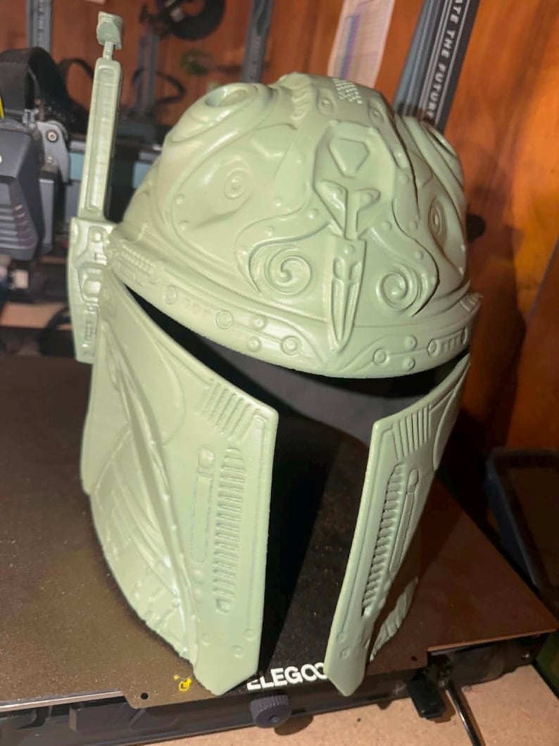 May include: A light green Mandalorian helmet with detailed features, including a T-shaped visor and antenna. The helmet is made of a robust material and has a complex design with various shapes and patterns. The helmet is a prop or costume piece.