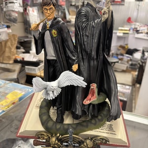 Może przedstawiać: Szczegółowa figurka przedstawiająca Harry'ego Pottera i Voldemorta. Harry trzyma różdżkę, a Voldemort ma na sobie czarną szatę. Obecna jest również biała sowa i zielony wąż z otwartą paszczą. Podstawą jest książka z napisem "Harry Potter".