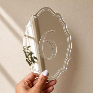 May include: A reflective, oval-shaped sign with a scalloped edge featuring the number 6 etched into the center. The surface is highly polished and mirror-like, showing reflections of the surrounding environment and light.