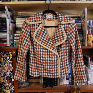 May include: A vintage houndstooth jacket in red, white, and blue, with a cream-colored trim and lining. The jacket is hanging on a black hanger. The label reads "Country Candy".