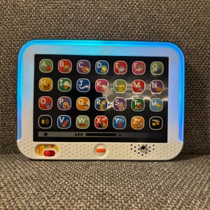 May include: A white and black educational tablet toy featuring a grid of colorful alphabet buttons from A to Z, each with a corresponding icon. The device has a glowing blue light border, a red slider switch, and a speaker grille on the front.