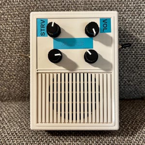 May include: A rectangular white guitar effects pedal featuring four black control knobs with white indicators. The faceplate includes blue labels reading STRV and VOL, with the letters AJC centered. The lower section has a vertical grille design.