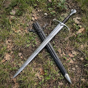 May include: A double-edged steel sword with a central fuller, a cross-shaped guard, and a dark grip. The sword is positioned diagonally across a black leather scabbard that features metal fittings and adjustable black leather straps.