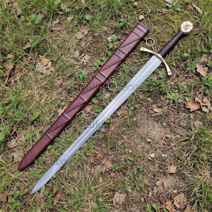 Peut inclure: Une épée en acier avec une poignée gainée de cuir marron foncé, une garde dorée et un pommeau orné d'un emblème de lion rouge. L'épée est accompagnée d'un fourreau en cuir marron foncé assorti avec des anneaux en métal doré.