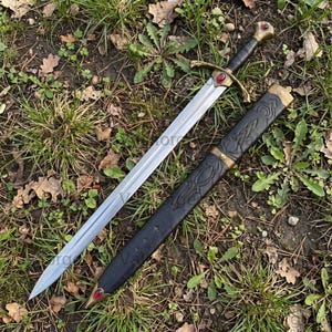 May include: A double-edged steel sword with a black leather-wrapped grip, a brass crossguard featuring a central red gemstone, and a matching pommel. The black scabbard has embossed patterns, brass fittings, and a red gemstone at the tip.
