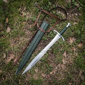 Puede incluir: Una espada de acero de doble filo con empuñadura verde oscuro y guarda plateada. Junto a ella hay una vaina verde oscuro a juego con una cresta central, acompañada de un arnés de cuero marrón con hebillas y correas de metal plateado.