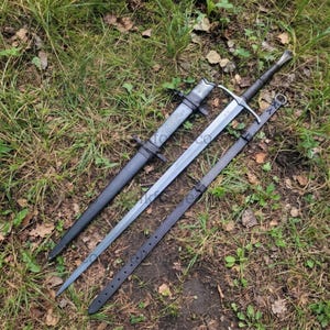 May include: A long steel sword with a crossguard and a dark handle, positioned next to a matching black scabbard and a separate black leather belt strap with metal eyelets, all resting on a patch of green grass and brown earth.
