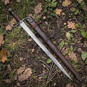 Puede incluir: Una espada de acero con hoja de doble filo, una guarda con un gavilán curvado y una empuñadura marrón texturizada. La espada va acompañada de una vaina de cuero marrón a juego con detalles de cuero trenzado cerca de la boca.