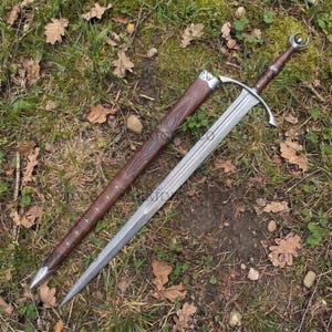 Op de afbeelding: Een zilverkleurig metalen zwaard met een lang, tweesnijdend lemmet, een pareerstang en een bruin handvat met een halvemaanvormige pommel. Daarnaast ligt een bijpassende bruine schede met decoratieve reliëfpatronen en kleine kruisaccenten.