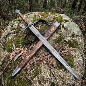 May include: A double-edged sword with a patterned Damascus steel blade, a brown wooden grip, and a crossguard. The matching wooden scabbard features gold-coloured fleur-de-lis emblems, intricate carvings, and metal fittings at the tip and throat.