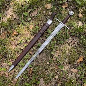 May include: A double-edged steel sword with a straight crossguard and a dark brown leather-wrapped grip, featuring a matching dark brown leather scabbard with a silver-toned metal chape and locket, positioned diagonally on a patch of grass.