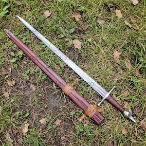 May include: A long, double-edged steel sword with a pointed tip and a central fuller. The hilt features a straight crossguard and a rounded pommel. Beside the blade lies a dark red scabbard with tan leather straps wrapped around the exterior.
