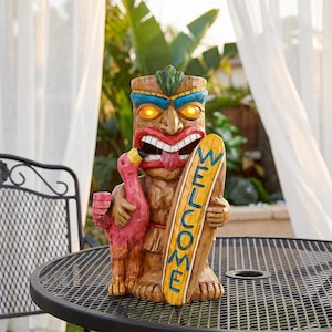 May include: A brown tiki statue with glowing yellow eyes, a red mouth, and a green topknot. The figure holds a pink flamingo in one hand and a yellow surfboard with the blue text WELCOME in the other, featuring a wood-carved texture throughout.