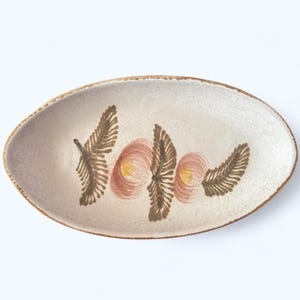 May include: An oval-shaped ceramic dish with a speckled off-white glaze and a brown rim. The interior features a hand-painted design of three dark brown fern-like leaves interspersed with two circular pink motifs containing yellow centers.