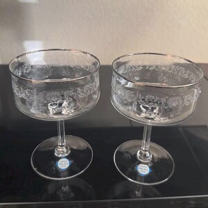 May include: Two clear champagne coupe glasses with silver rims and floral etched designs. The glasses have a stem and a round base. The glasses are on a black surface.