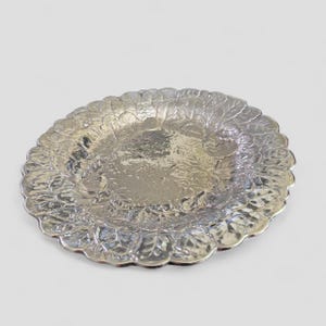 May include: A round silver-toned metal plate featuring a detailed embossed pattern of overlapping leaves covering the entire surface. The plate has a scalloped edge and a reflective, polished finish that catches the light across its textured design.