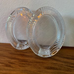 May include: Two clear glass candle holders with an oval shape and a textured, diagonal ribbed pattern. Each piece features a smooth, recessed circular center designed to hold a single candle, with a thick, molded glass frame surrounding it.
