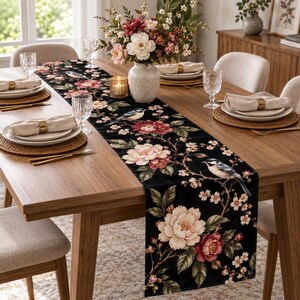May include: A black table runner featuring a botanical print of large pink and white peony flowers, green leaves, and small birds perched on thin branches. The runner is laid out on a wooden dining table set with plates, napkins, and glassware.