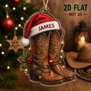 May include: A 2D flat acrylic Christmas ornament shaped like a pair of brown leather cowboy boots with intricate stitching, adorned with a red Santa hat featuring the name JAMES, green holly leaves with red berries, and string lights.