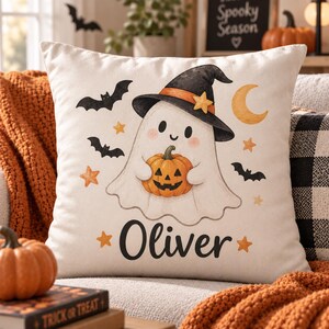 May include: A square off-white throw pillow featuring a central illustration of a white ghost wearing a black witch hat with an orange band and yellow star. The ghost holds an orange jack-o'-lantern. The name Oliver is printed in black below.