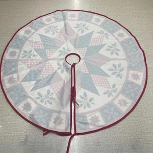 May include: A circular Christmas tree skirt featuring a central star pattern in shades of light blue, pink, and white. The border displays a repeating square motif with holly leaves and star shapes, finished with a dark red fabric trim and ties.