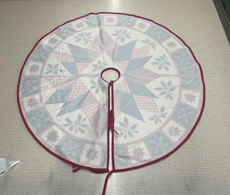 May include: A circular Christmas tree skirt featuring a central star pattern in shades of light blue, pink, and white. The border displays a repeating square motif with holly leaves and star shapes, finished with a dark red fabric trim and ties.