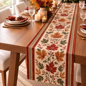 May include: A rectangular table runner featuring a central pattern of autumn maple leaves in shades of red, orange, and green, bordered by vertical stripes in deep red and olive green on a cream-colored fabric base.