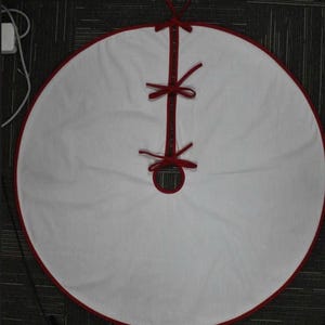 May include: A circular white Christmas tree skirt featuring a solid red trim along the outer edge and the center opening. The skirt includes a straight slit for placement around a tree base, secured by three red fabric ties spaced along the opening.