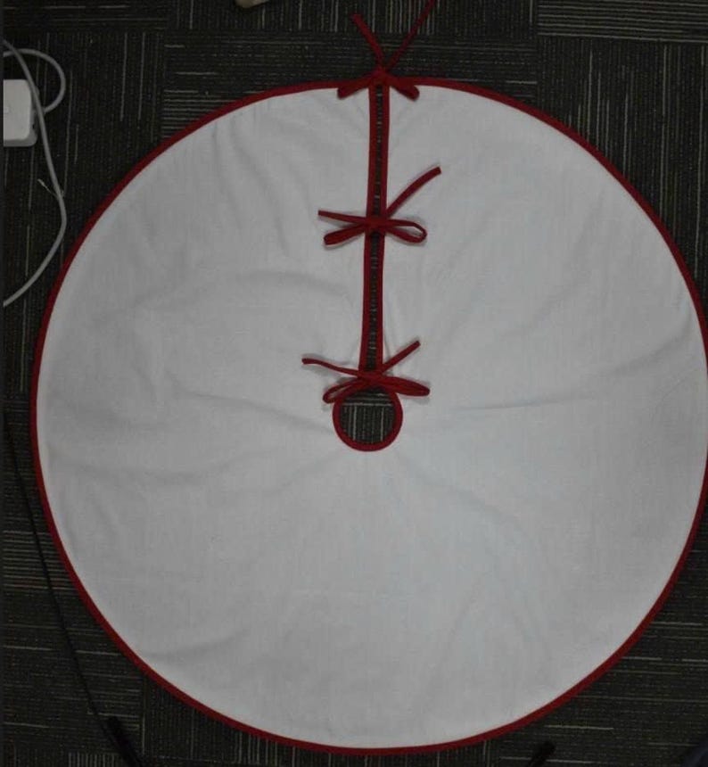 May include: A circular white Christmas tree skirt featuring a solid red trim along the outer edge and the center opening. The skirt includes a straight slit for placement around a tree base, secured by three red fabric ties spaced along the opening.