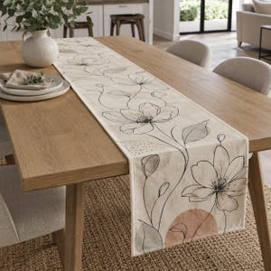 May include: A long rectangular table runner featuring a cream-colored fabric base with a black line-art floral pattern. The design includes delicate stems, leaves, and large blossoms, accented by soft, muted terracotta and sage green color washes.