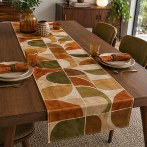 May include: A rectangular table runner featuring a geometric pattern of semicircles and squares in shades of burnt orange, olive green, and cream. The runner is laid across a dark wood dining table set with plates, orange napkins, and glassware.
