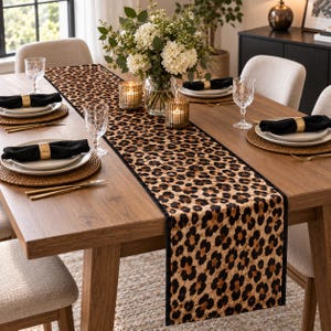 May include: A rectangular table runner featuring a brown and black leopard print pattern with a solid black border. The runner is centered on a wooden dining table set with white plates, black cloth napkins with gold rings, and clear glasses.