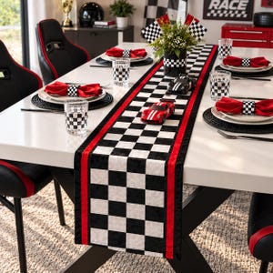 May include: A dining table set with a black and white checkered table runner featuring red borders, matching checkered napkins with napkin rings, checkered drinking glasses, and two toy race cars placed along the center of the table runner.